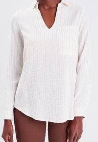 Chemise blanche à manches longues avec un col en V, poche avant, rayures verticales et petits accents à pois, fabriquée en tissu léger.