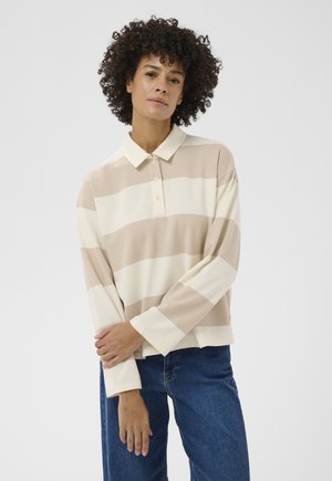 Frau mit lockigem Haar, die ein beigefarbenes und weiß gestreiftes langärmeliges Poloshirt und blaue Jeans trägt, steht mit verschränkten Armen vor einem weißen Hintergrund.