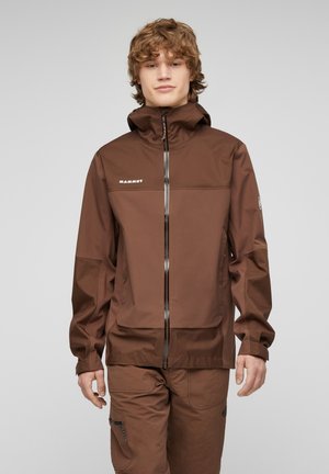 Jong persoon met krullend haar, gekleed in een bruine Mammut-jas en bijpassende broek tegen een effen grijze achtergrond.