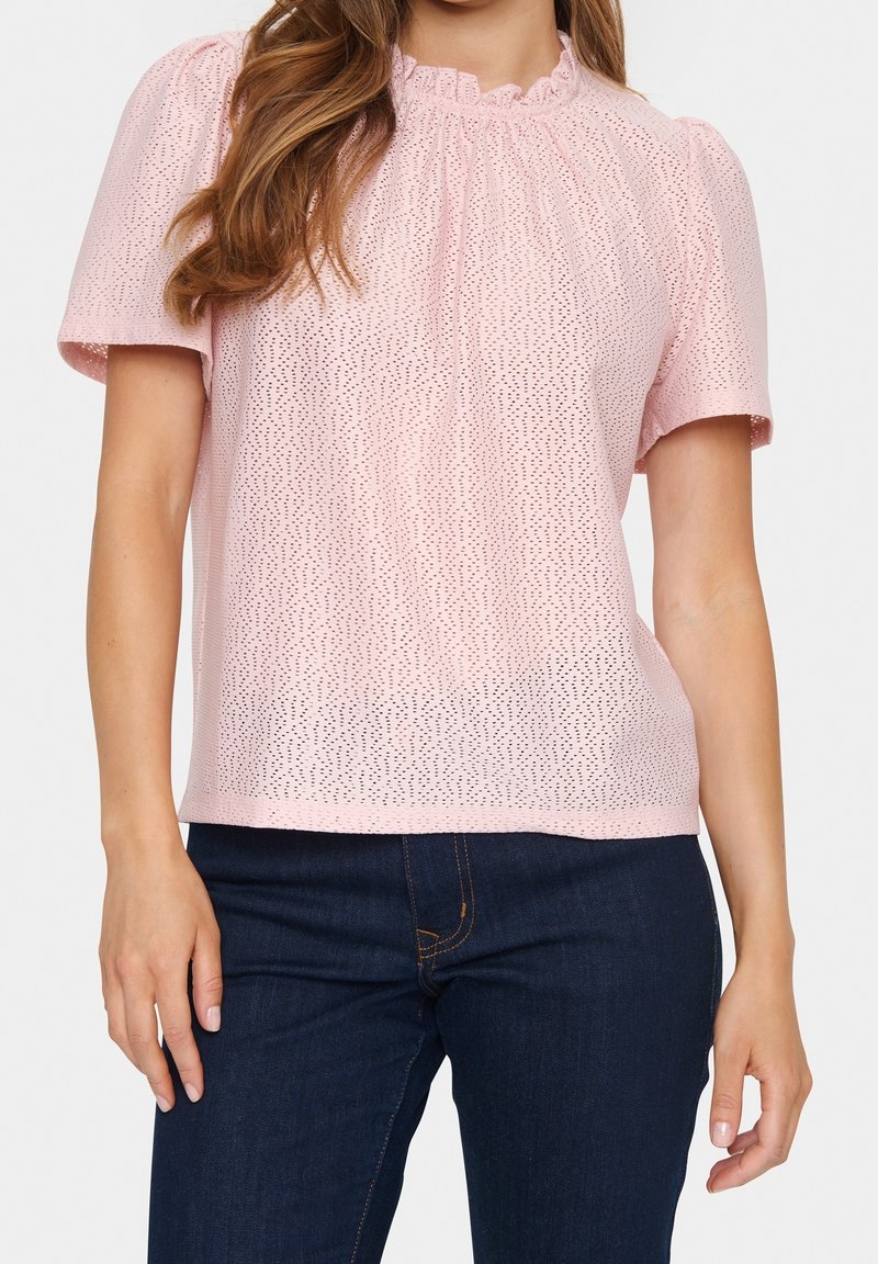 Femme portant un chemisier rose clair à manches courtes avec œillets et col froncé, ainsi qu'un jean bleu foncé sur fond blanc.