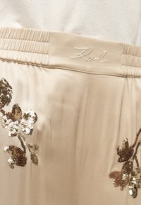 Bež satenske hlače z elastičnim pasom. Imajo vezenje cvetnih oblik v sekvinskem okrasju, z zapletenimi podrobnostmi v rjavi in srebrni barvi.