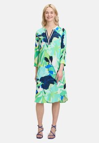 Sukienka w kwiatowy wzór z dekoltem w szpic, rękawami 3/4 i luźnym krojem. Przeważający jasny zielony kolor, z akcentami w ciemnym niebieskim i teal.