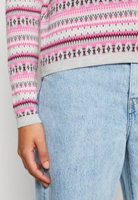 Pull en laine gris avec des motifs géométriques roses, noirs et blancs ; associé à un jean en denim bleu clair. Gros plan sur le bas de la manche et la taille.