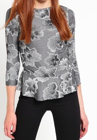 Woman wearing a grey peplum top with black and white floral patterns and three-quarter sleeves, paired with fitted black pants.