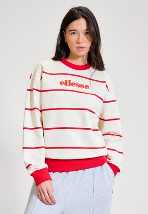 Frau mit langen dunklen Haaren, die einen weißen Pullover mit roten Querstreifen und dem "ellesse"-Logo trägt, kombiniert mit hellgrauen Hosen, steht vor weißem Hintergrund.