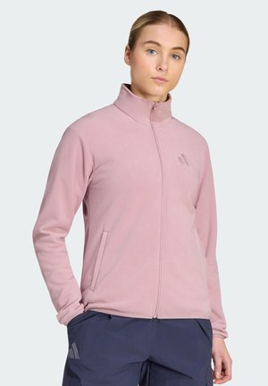 Vrouw met blond haar in een roze jas met rits en donkerblauwe broek, staand met één hand in de zak tegen een effen lichte achtergrond.
