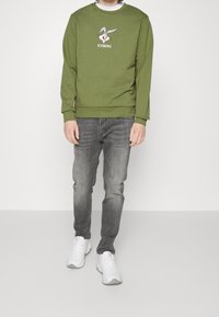 Felpa verde in cotone con grafica di Bugs Bunny, testo "ICEBERG", jeans slim-fit grigi e sneakers bianche. Design semplice, stile confortevole.