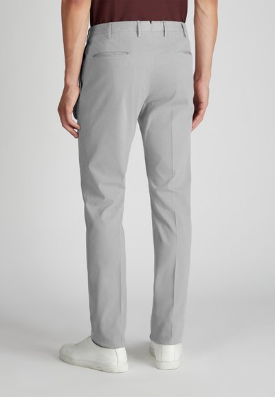 Mężczyzna w jasnoszarych, dopasowanych spodniach i białych sneakersach, stojący z luzem przy boku na jednolitym tle.