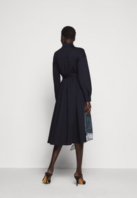 Abito lungo a maniche, di colore blu navy, con vita cintata, gonna plissettata e orlo decorato. Tessuto liscio con colletto strutturato. Tacchi visibili.