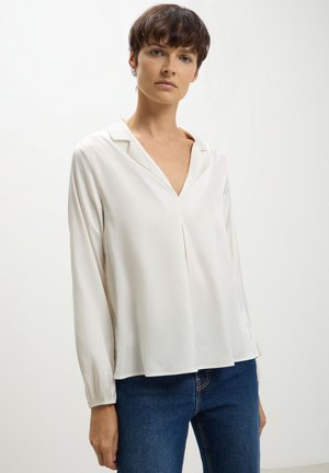 Blusa bianca a maniche lunghe con scollo a V e vestibilità rilassata. Realizzata in tessuto morbido, presenta un dettaglio plissettato sul davanti e maniche arricciate.