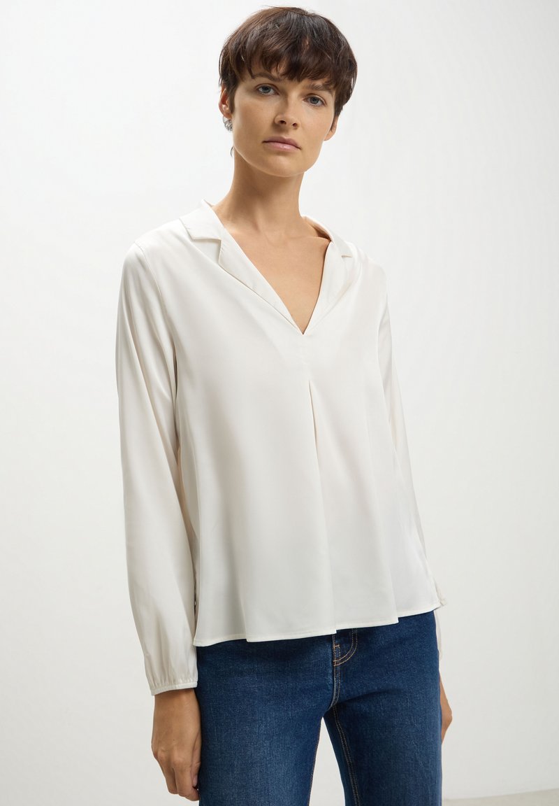 Blusa bianca a maniche lunghe con scollo a V e vestibilità rilassata. Realizzata in tessuto morbido, presenta un dettaglio plissettato sul davanti e maniche arricciate.