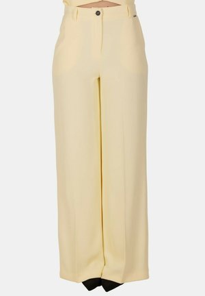 Pantaloni larghi color crema a vita alta con chiusura frontale con bottone e zip, tasche laterali, indossati con scarpe nere con punta appuntita.