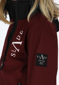 Maroon jacket with a black collar; features white logo on the chest and a black patch on the sleeve with brand details. Zippered pockets visible.