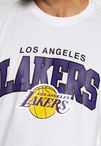 Primer plano de una camiseta blanca con el logo de los Los Angeles Lakers que presenta texto púrpura y un gráfico de baloncesto amarillo.