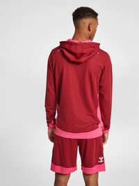 Röd idrottströja med rosa detaljer och dragsko i huvan, i kombination med matchande röda shorts med rosa kant. Tillverkad av texturerad tyg.