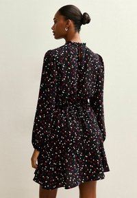 Schwarzes Kleid mit einem Punktenmuster in Pink und Weiß. Hat lange Ärmel, einen Bindegürtel und ein Ausschnitt-Detail im Rücken. Ausgestellter Saum.