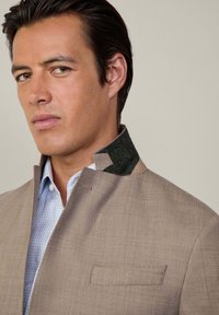 Veste beige avec un col haut, présentant des accents vert foncé, des épaules structurées et un tissu texturé. Superposée sur une chemise à carreaux bleu clair.
