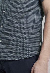 Dettaglio di una persona che indossa una maglietta nera a maniche corte con un piccolo motivo geometrico bianco e verde e pantaloni scuri.