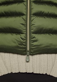 Olivengrøn pufferjakke med lynlås foran, der har en struktureret beige strikket kant og et glat sort underlag.