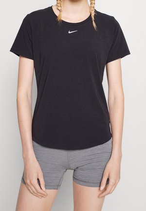 Persona vestida con una camiseta negra de Nike y pantalones cortos ajustados grises, de pie con las manos relajadas a los lados contra un fondo neutro.