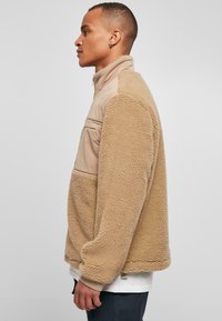 Pullover in pile beige con un pannello frontale in nylon color sabbia. Presenta un colletto alto e polsini elasticizzati, corpo testurizzato e una tasca sul petto.