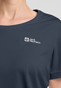 Una maglietta a maniche corte di colore blu navy, realizzata in tessuto liscio, con un logo bianco di Jack Wolfskin sul petto e un colletto rotondo.