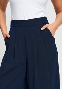 Morske široke hlače s visokim strukom, nabranim prednjim dijelom i bočnim džepovima, izrađene od glatkog, laganog materijala.