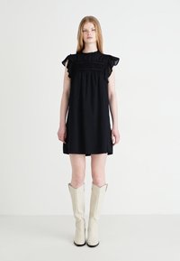 Μαύρο, κοντό φόρεμα με φουσκωτά μανίκια και σμιλευμένο μπούστο; υφασμάτινη υφή με διακριτό σχέδιο, συνδυασμένο με λευκά μποτάκια μέχρι το γόνατο.