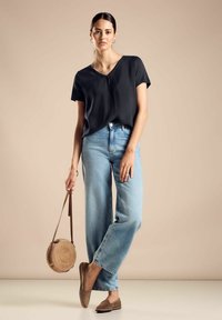 Sort kortærmet v-hals top, lyseblå brede jeans, brune flade sko, holder en rund vævet taske med læderrem.