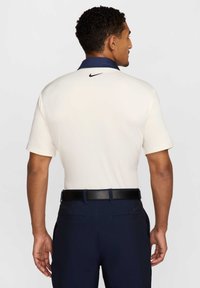 Kremowa koszulka polo z krótkimi rękawami i granatowym kołnierzykiem; posiada małe czarne logo Nike z tyłu. W zestawie z ciemnogranatowymi spodniami i czarnym paskiem.
