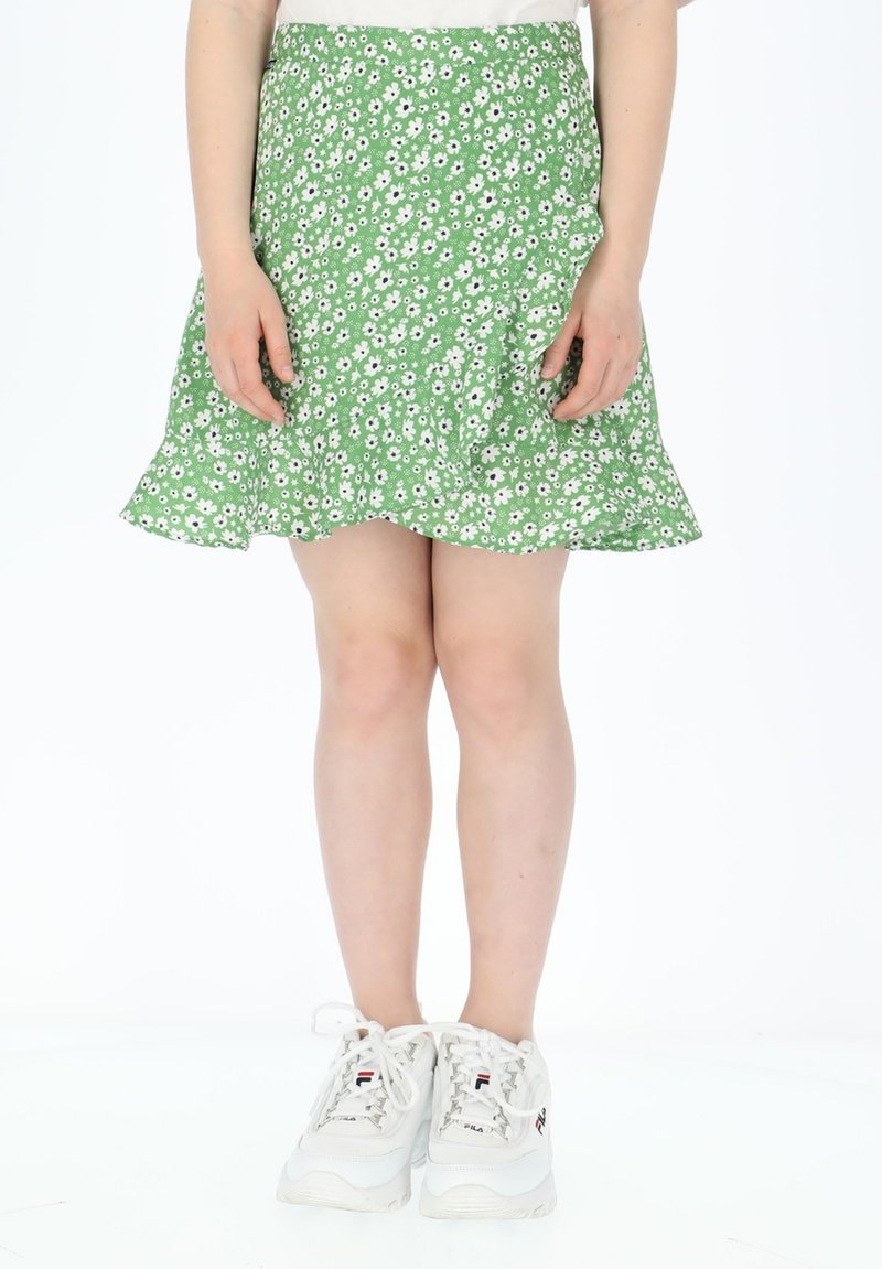 Grüner Blumenrock mit weißen Gänseblümchen, ausgestelltem Saum und leichtem Stoff. Getragen mit weißen Sneakers mit roten und blauen Akzenten.