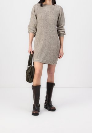 Frau in einem beigen, langärmligen Strickkleid, die eine kleine grüne Handtasche hält und hohe dunkelbraune Lederstiefel trägt.