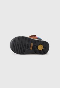 Semelle de chaussure multicolore présentant un fond en caoutchouc noir texturé avec des rainures, un logo doré et une tige en daim marron avec des accents.