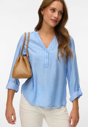 Femme portant un chemisier à rayures bleu clair et un pantalon blanc, avec un sac à main en cuir marron clair sur l'épaule, regardant sur le côté et souriant doucement.