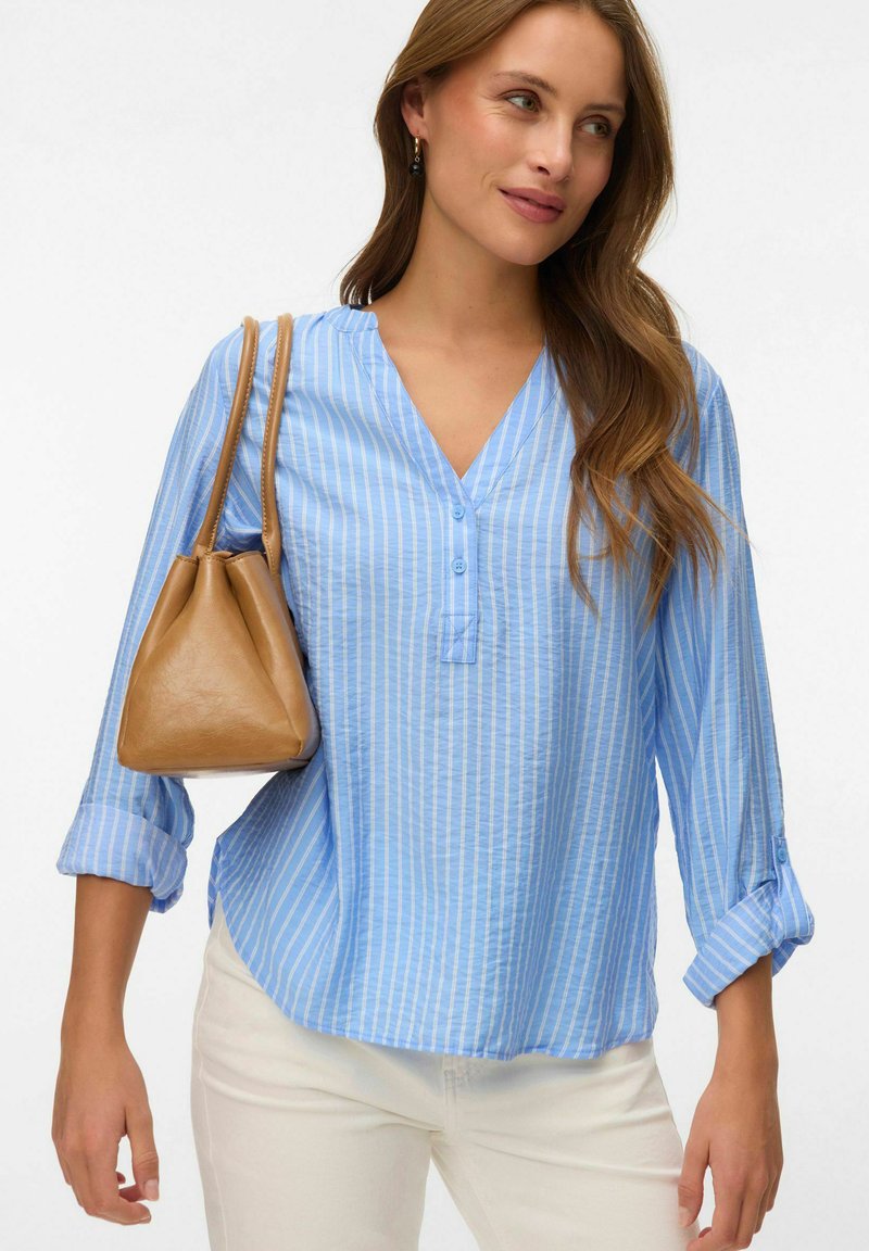 Femme portant un chemisier à rayures bleu clair et un pantalon blanc, avec un sac à main en cuir marron clair sur l'épaule, regardant sur le côté et souriant doucement.