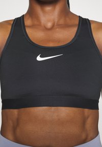 Black sports bra with a scoop neckline, thick underband, and white Nike swoosh logo on the front, made from stretchy, smooth fabric.