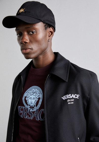 Sort zip-up jakke over en maroon t-shirt med et blåt Versace grafisk motiv af Medusa. Jakken har hvid broderet tekst og krave.