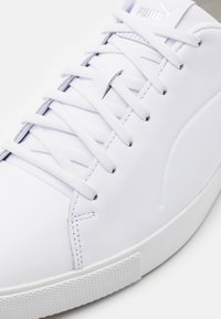 Vit lädersneaker med platta snören, texturerad gummisula och subtil logotypdetalj. Slät yta och minimalistisk design.