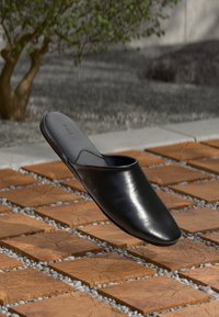 Sapato mule preto de couro, flutuando acima de ladrilhos de madeira quadrados, com seixos, uma pequena árvore e solo de pedras ao fundo embaçado.