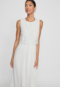Hvit kjole med blondeoverlegg med blomsterdesign, ermeløs fasong og en flytende skjørt. Glatt tekstur og elegant silhuett.