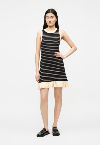 Γυναίκα που στέκεται με το χέρι στη μέση, φορώντας ένα μίντι αμάνικο φόρεμα με μαύρες και μπεζ ρίγες, με βολάν στο τελείωμα και μαύρα παπούτσια χωρίς κορδόνια.