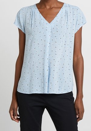 Jasnoniebieska bluzka z dekoltem w serek, z małym ciemnym abstrakcyjnym wzorem, krótkie rękawy, noszona z czarnymi spodniami.