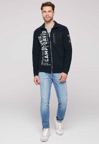Giacca zip-up blu navy con dettagli in bianco, tasca frontale e polsini a costine, abbinata a jeans azzurri e sneakers bianche.