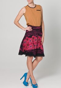 Top senza maniche marrone con rifiniture nere, abbinato a una gonna nera con motivi floreali rosa e scritte a mano, completato da tacchi blu.