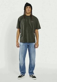 Dark green short-sleeve t-shirt with a left chest logo, paired with light blue jeans and black sneakers with white soles.