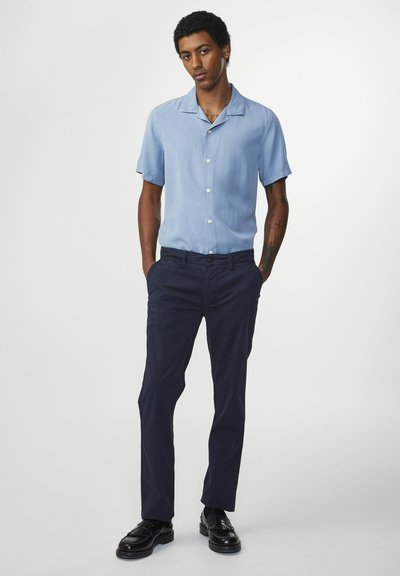 Camicia azzurro chiaro a maniche corte con colletto a camicia, chiusura a bottoni, abbinata a pantaloni blu navy e scarpe di pelle nere. Tessuto dalla superficie liscia.