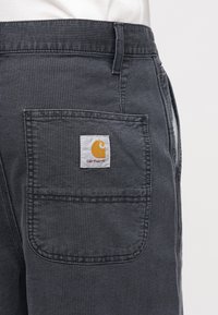 Nærbilde av lomme på mørkegrå kordfløyelbukser med Carhartt-logo påsydd, brukt over en hvit skjorte.