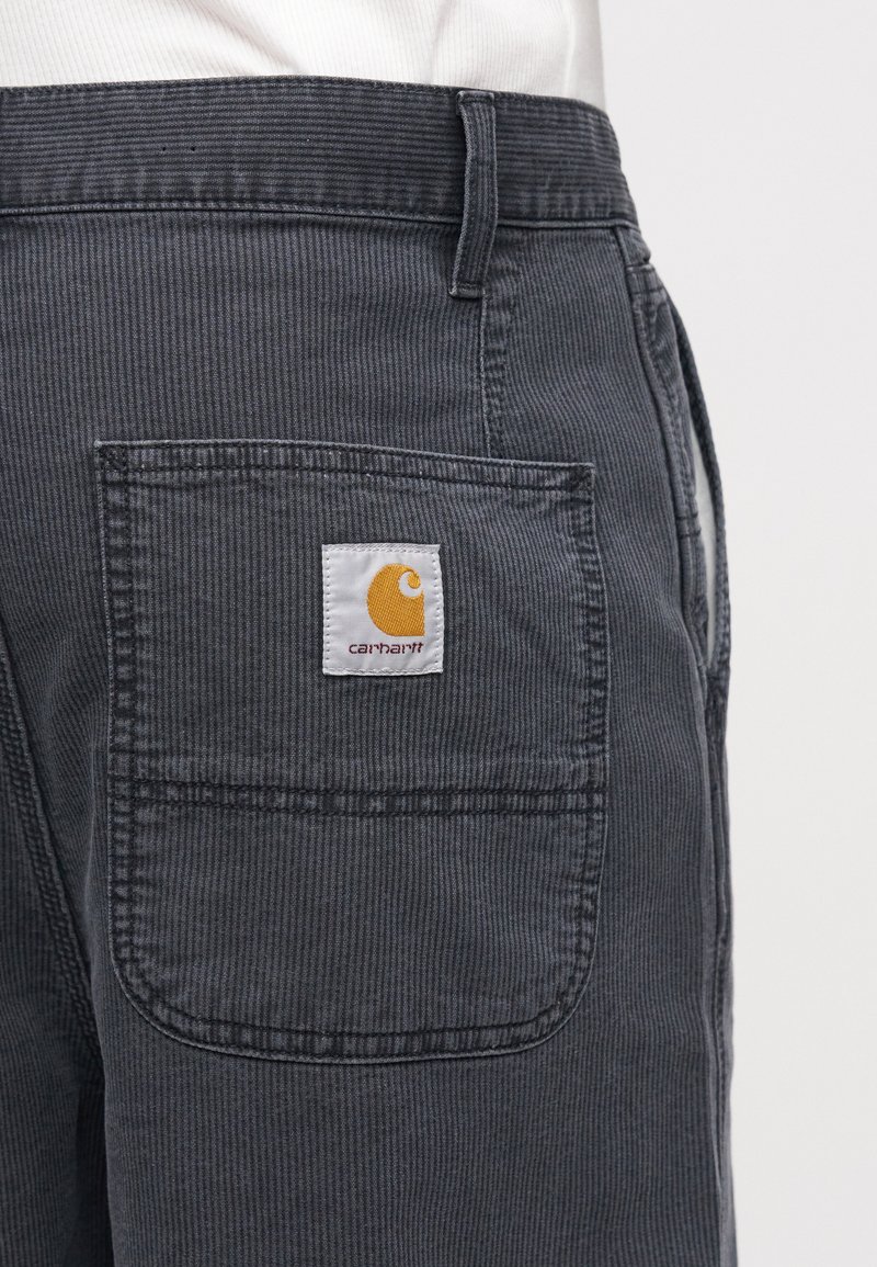 Primo piano della tasca di pantaloni in velluto a coste grigio scuro con toppa con il logo Carhartt indossati sopra una camicia bianca.