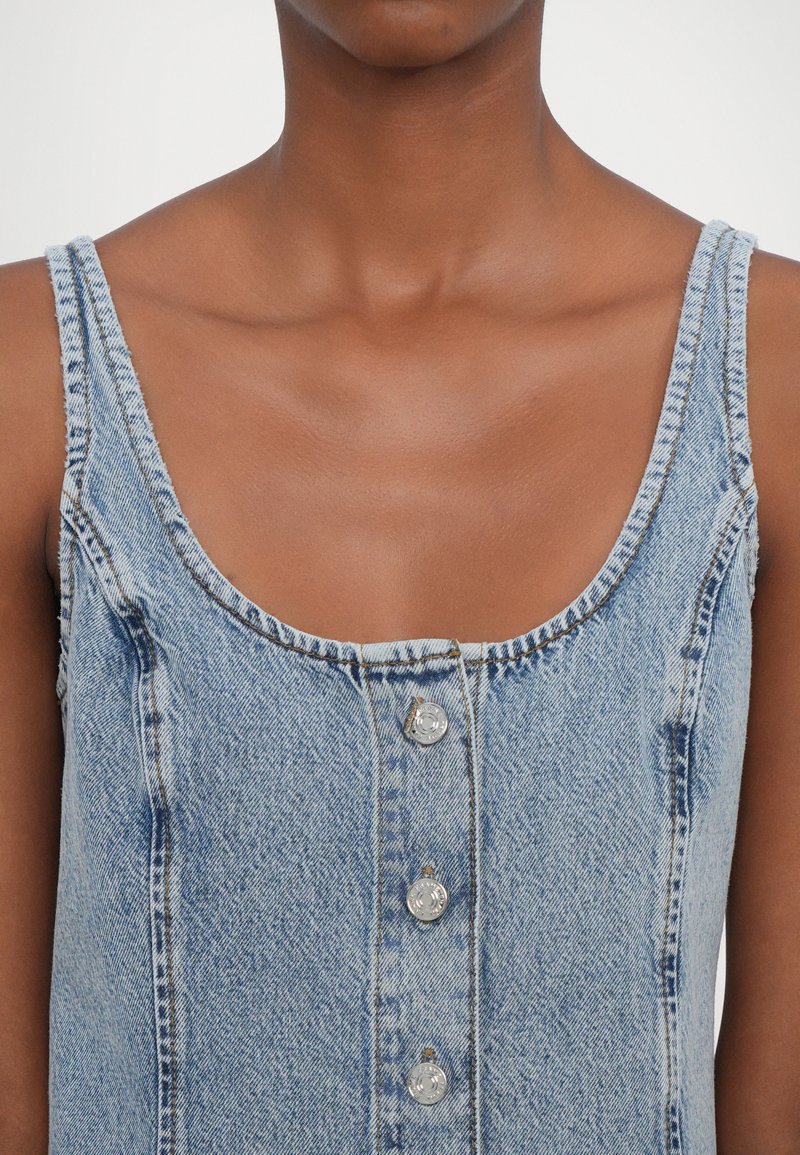 Helles blaues Denim-Kleid mit rundem Halsausschnitt, breiten Trägern und vorderer Knopfleiste mit fünf silbernen Knöpfen. Glatte Textur.
