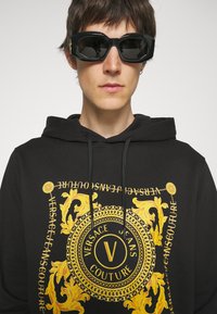 Felpa nera con un grafico barocco dorato caratterizzato da un design circolare, testo e dettagli accentuati. Modello che indossa occhiali da sole neri oversize.